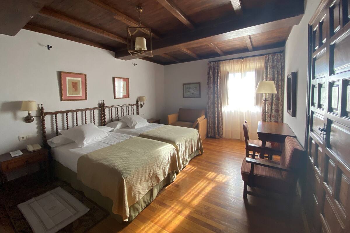 A room with two beds, a wooden ceiling, a dining table with chairs, and a window with curtains.
