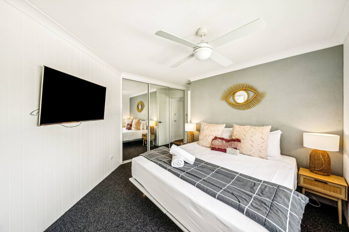 A hotel room with a bed, a television, a ceiling fan, a mirror, and a nightstand with a lamp.
