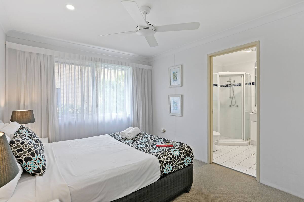 A hotel room with a bed, a ceiling fan, a shower, and a door leading to the bathroom.
