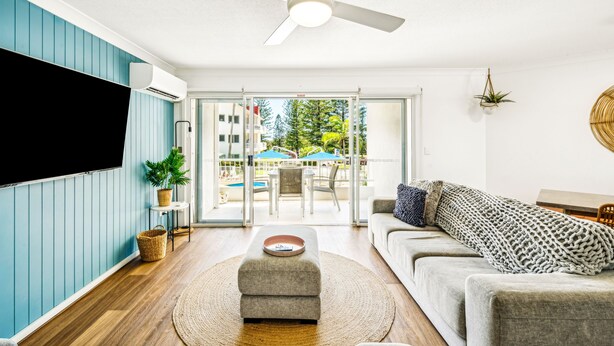A living room with a flat-screen TV, a ceiling fan, a sofa, a coffee table, and a view of an outdoor pool area. - Le Beach Apartments (Burleigh Heads)