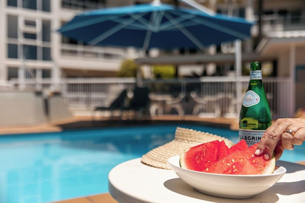 A poolside setting with a bowl of watermelon slices, a bottle of beer, and a blue umbrella.
