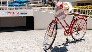 A red bicycle with a basket and a hat rack, parked on a concrete surface with a white railing and palm trees in the background. - Le Beach Apartments (Burleigh Heads)