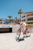 A red bicycle with a basket and a hat rack, parked on a concrete surface with a white railing and palm trees in the background.