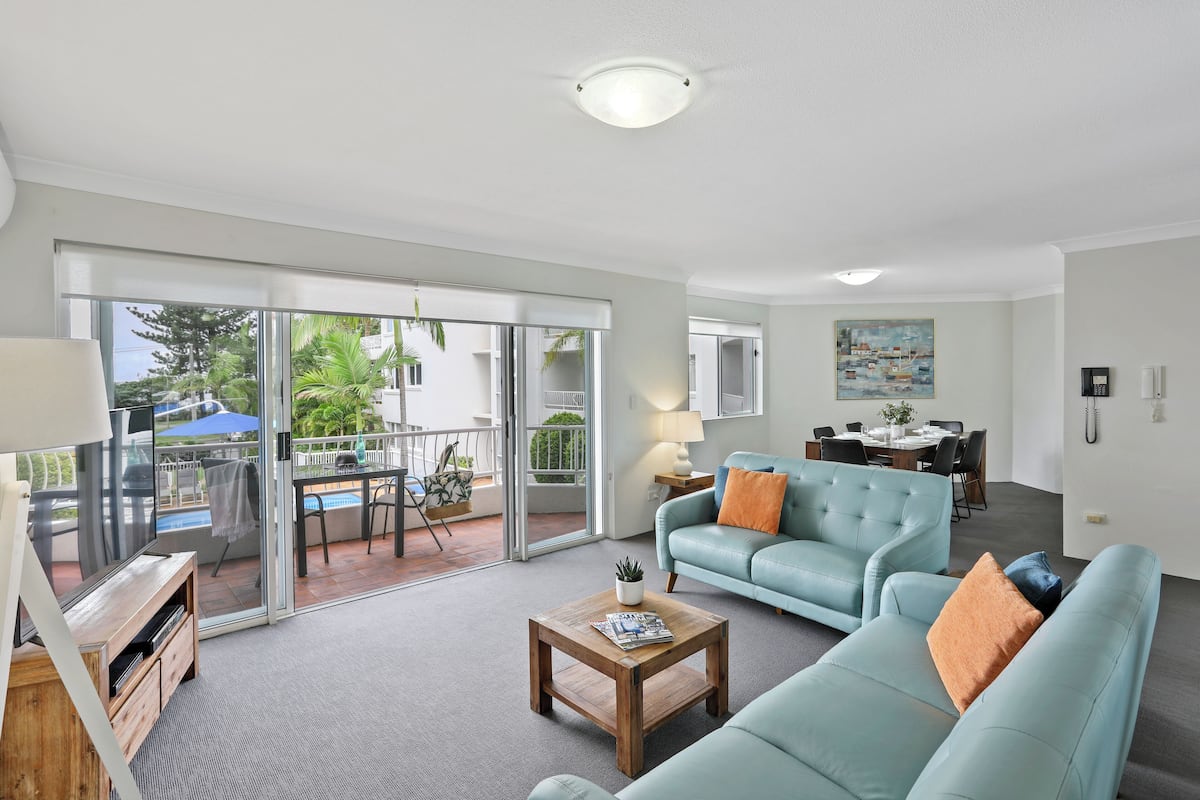 A spacious living area with a sofa, coffee table, and a dining table with chairs. There is a sliding glass door leading to an outdoor patio with a pool and umbrellas.