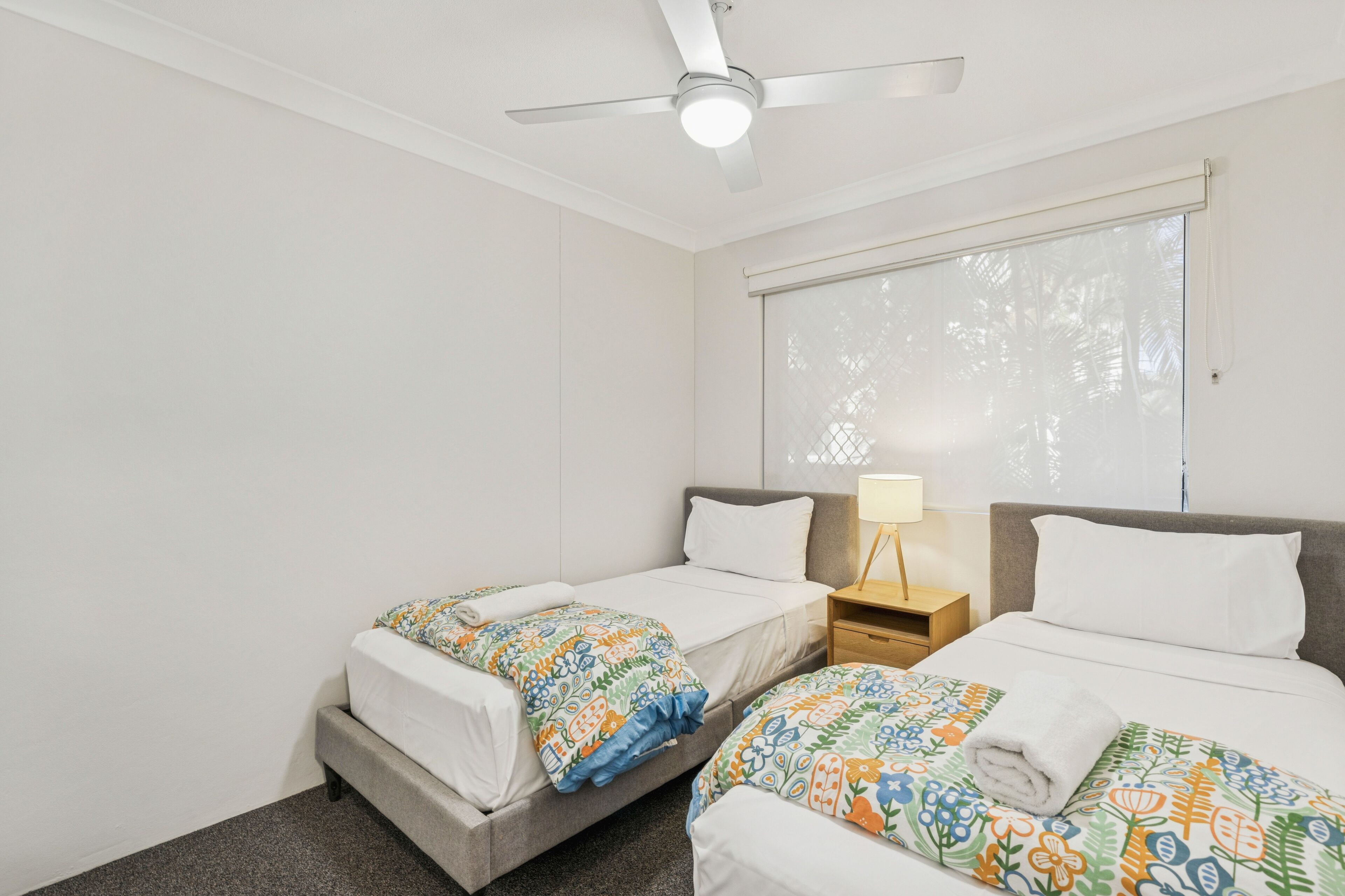 A hotel room with two single beds, a ceiling fan, a window with blinds, and a bedside table with a lamp.