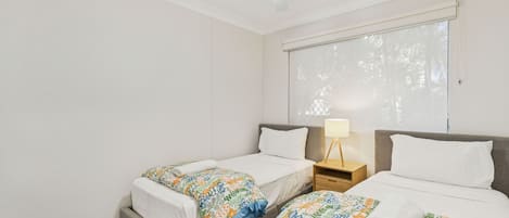 A hotel room with two single beds, a ceiling fan, a window with blinds, and a bedside table with a lamp.