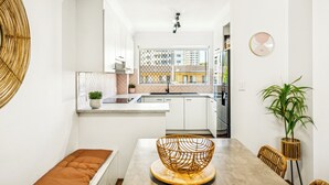 A modern kitchen with white cabinets, a large window, and a dining area with a wooden table and bench. - Le Beach Apartments (Burleigh Heads)