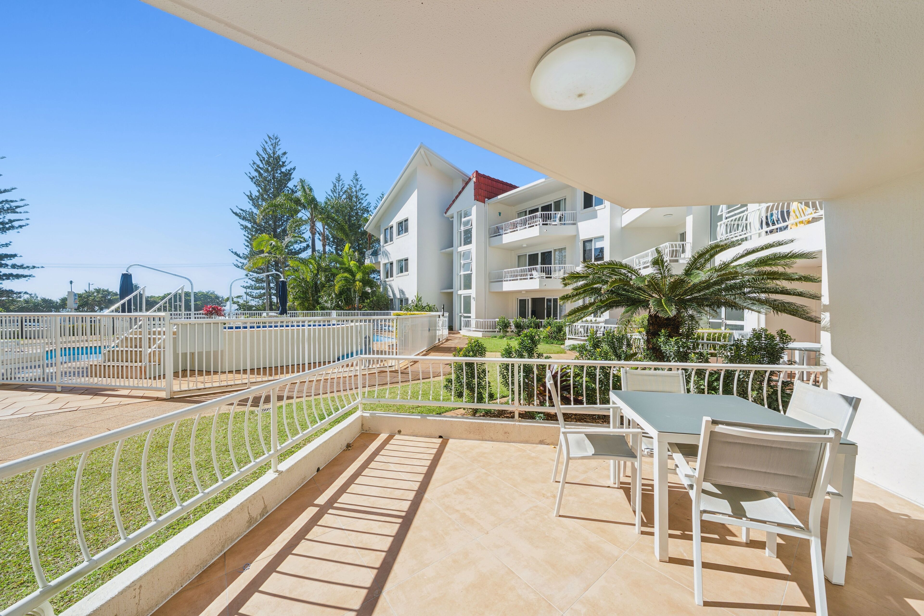 A balcony with a table and chairs, a pool, and a building with multiple balconies in the background.