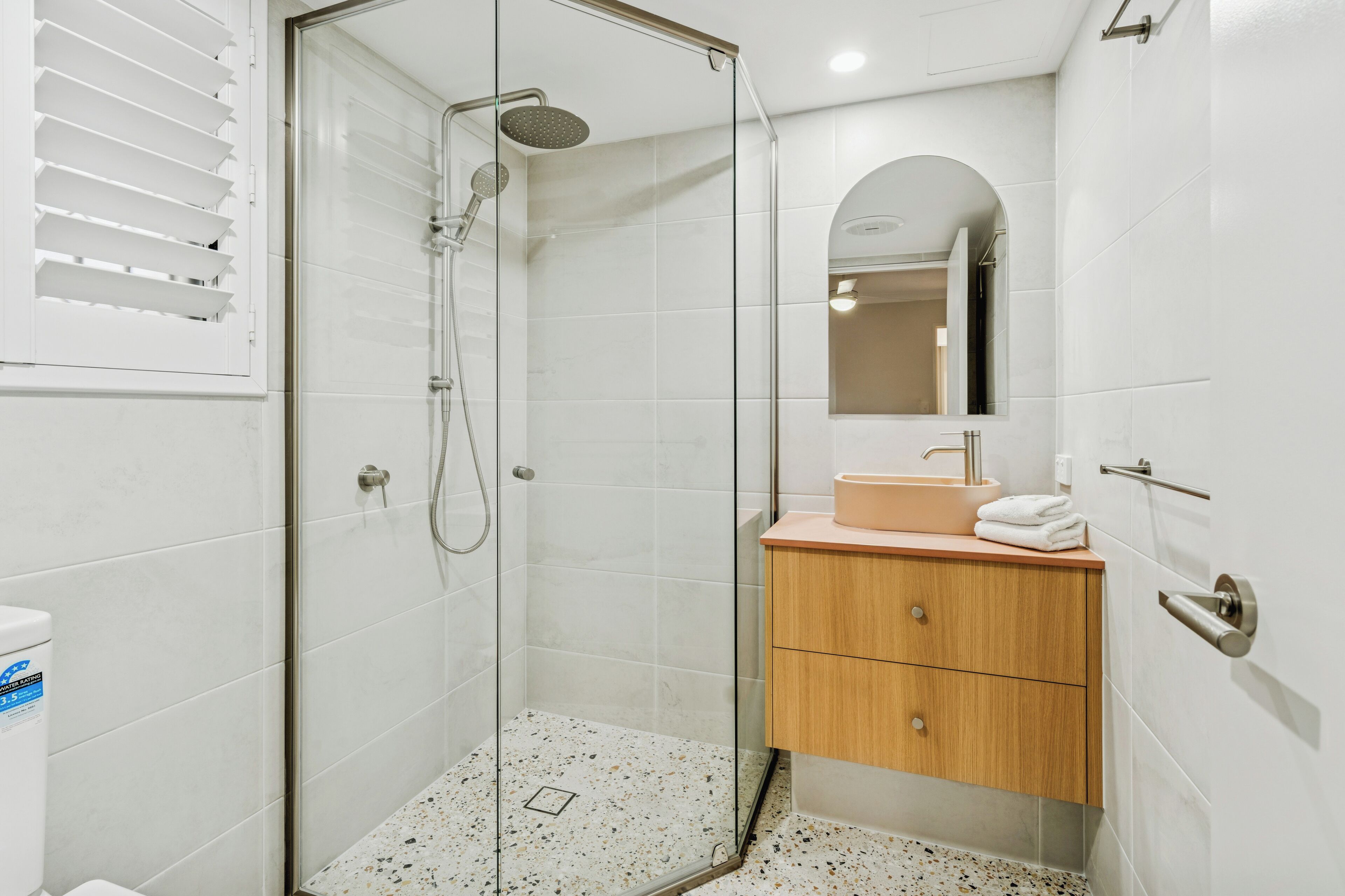 A modern bathroom with a glass-enclosed shower, a wooden vanity, and a white tiled floor.