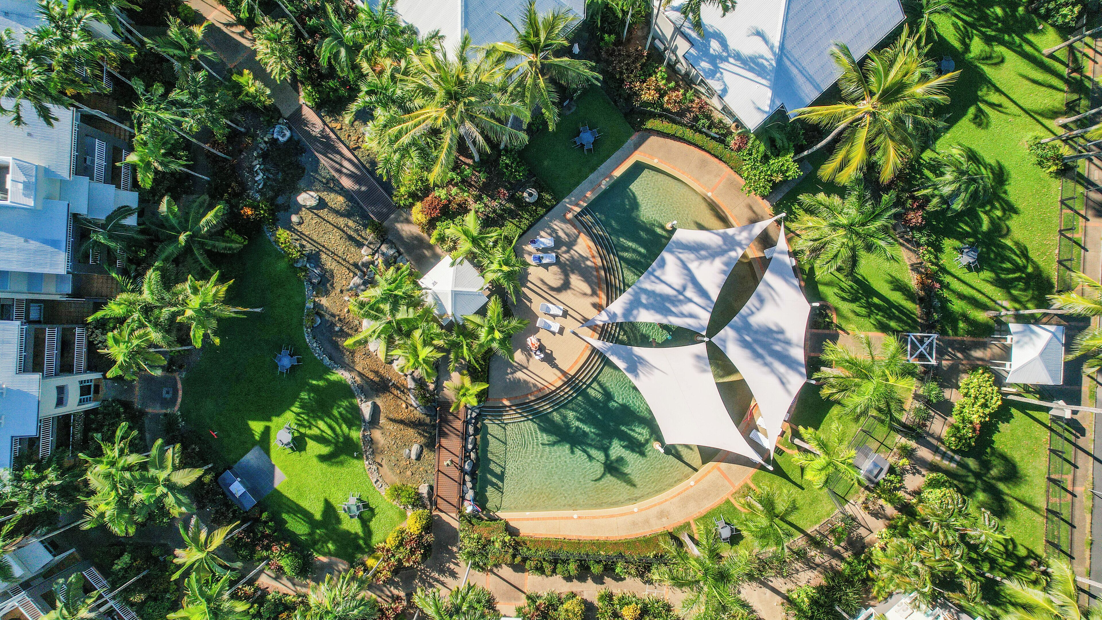 An aerial view of a resort with a central pool, surrounded by palm trees and shaded areas.