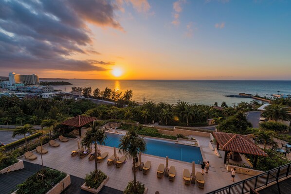 A resort with a swimming pool, lounge chairs, and a view of the sunset over the sea.