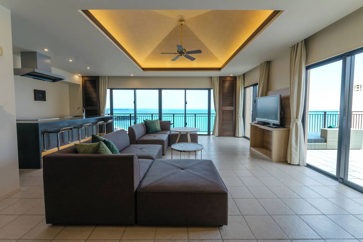 Een moderne woonkamer met een grote hoekbank, een kleine ronde tafel en een televisie op een houten dressoir. De kamer heeft een plafondventilator, een groot raam met uitzicht op zee en een balkon met tuinmeubilair.