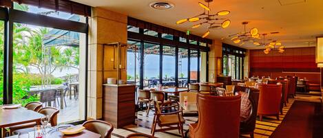 Un restaurante con vista al océano, con mesas y sillas de madera, una zona de bar y amplias ventanas.