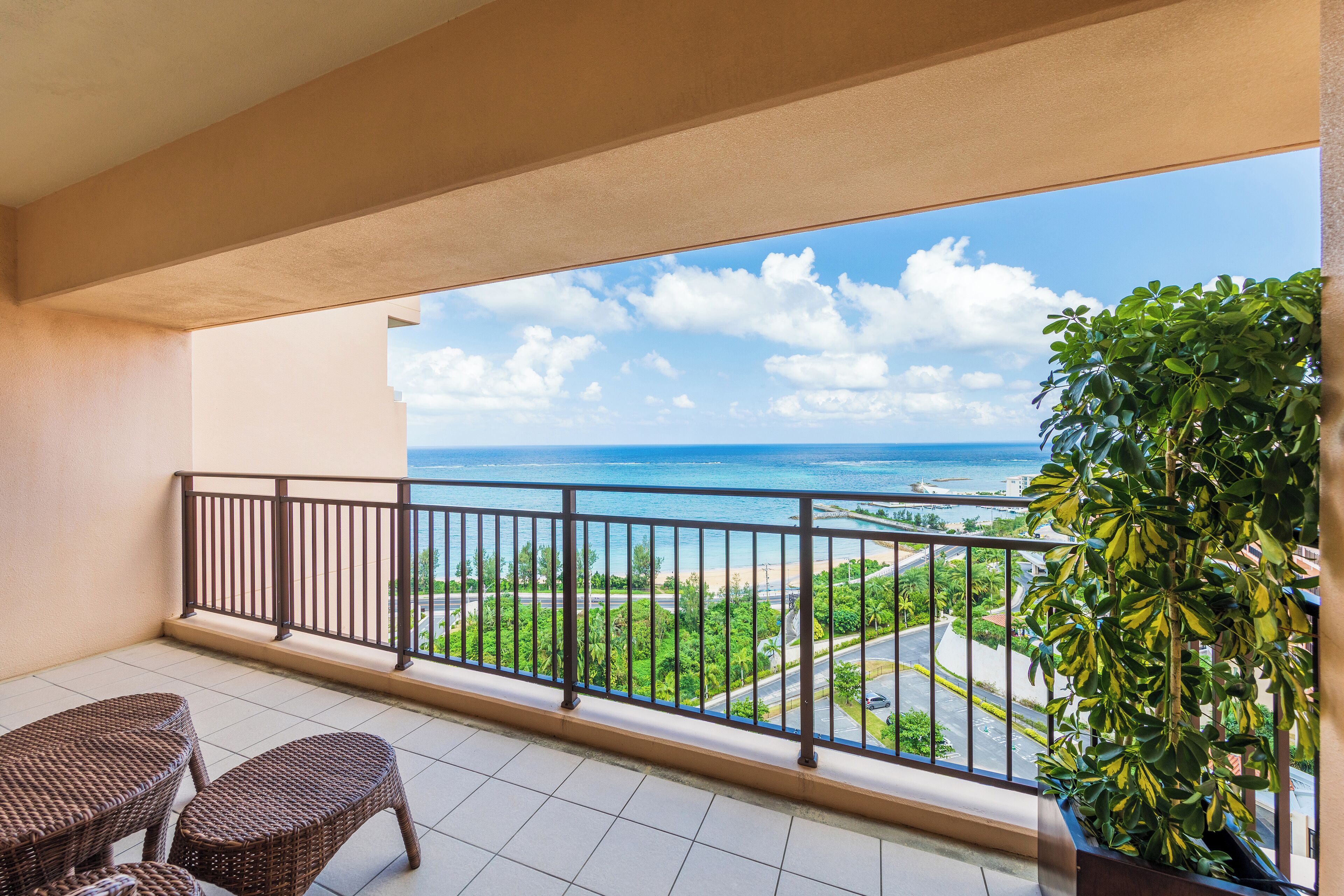 A balcony with wicker furniture, a potted plant, and a view of the ocean and greenery.