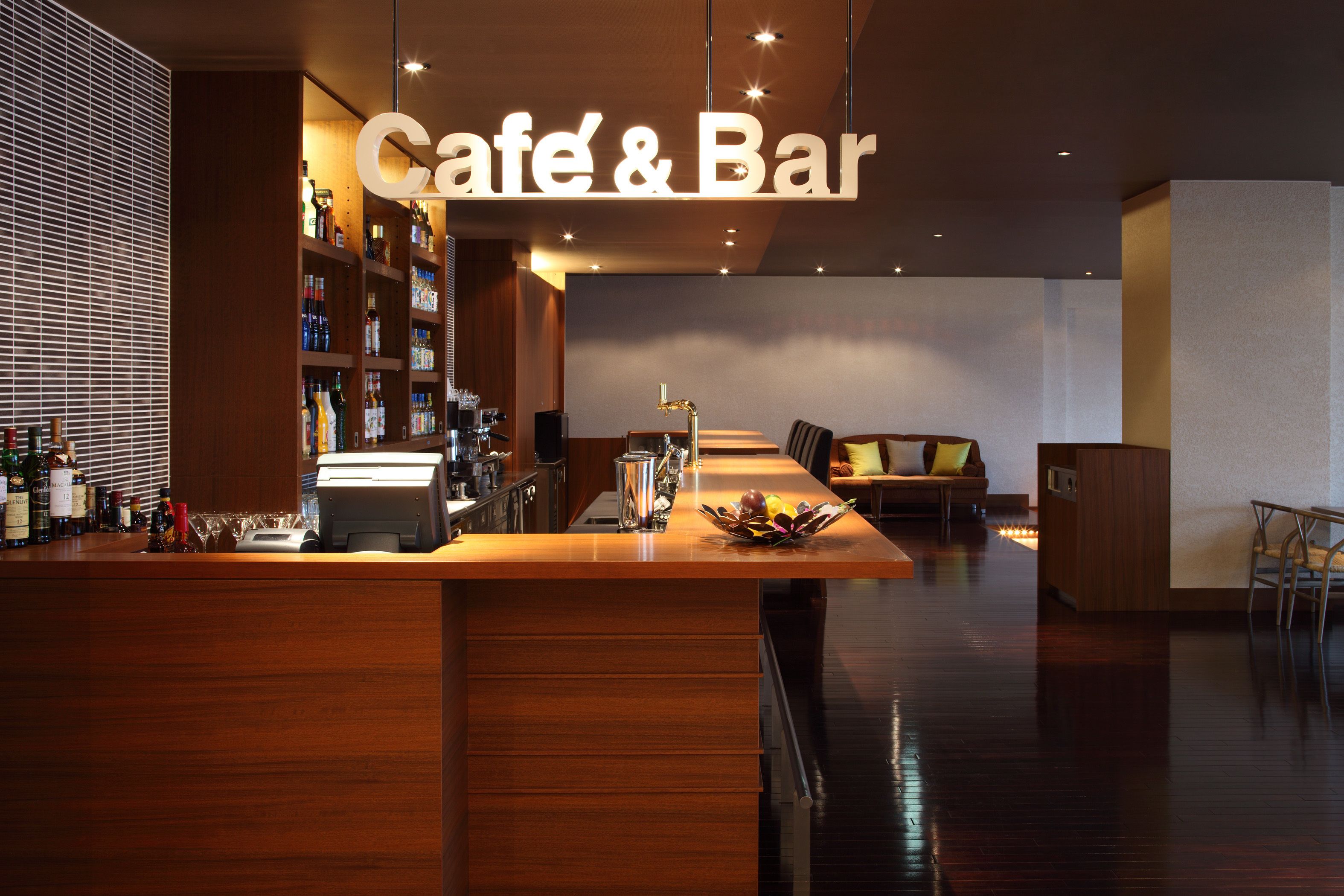 A modern café and bar interior with a long wooden counter, shelves of beverages, and a seating area with a sofa and chairs.