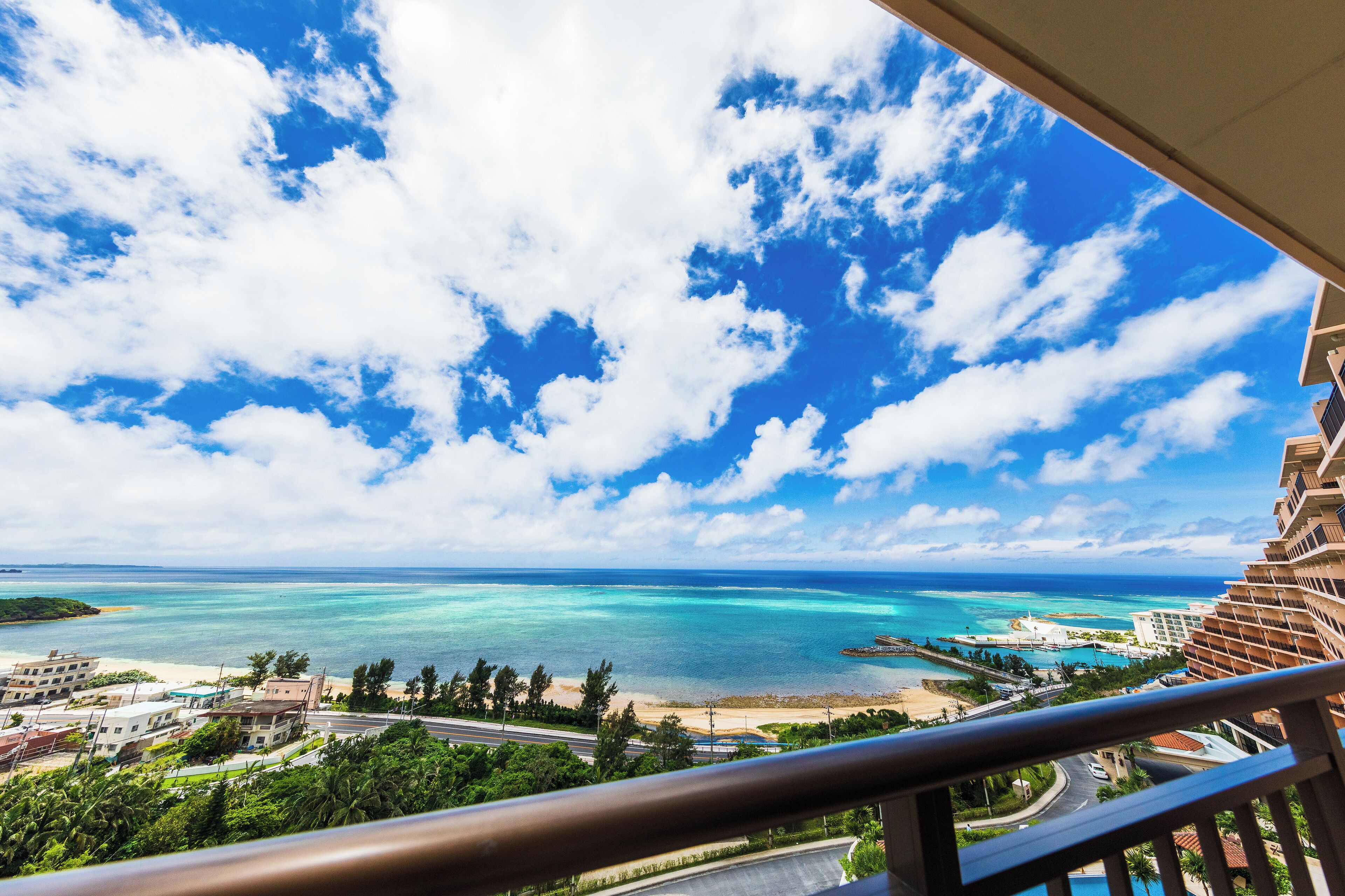 A coastal view from a balcony overlooking turquoise waters, a beach, and a resort area.