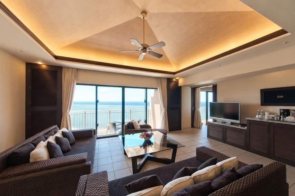 Een moderne woonkamer met een plafondventilator, grote ramen, een flatscreen-tv en een balkon met uitzicht op zee.