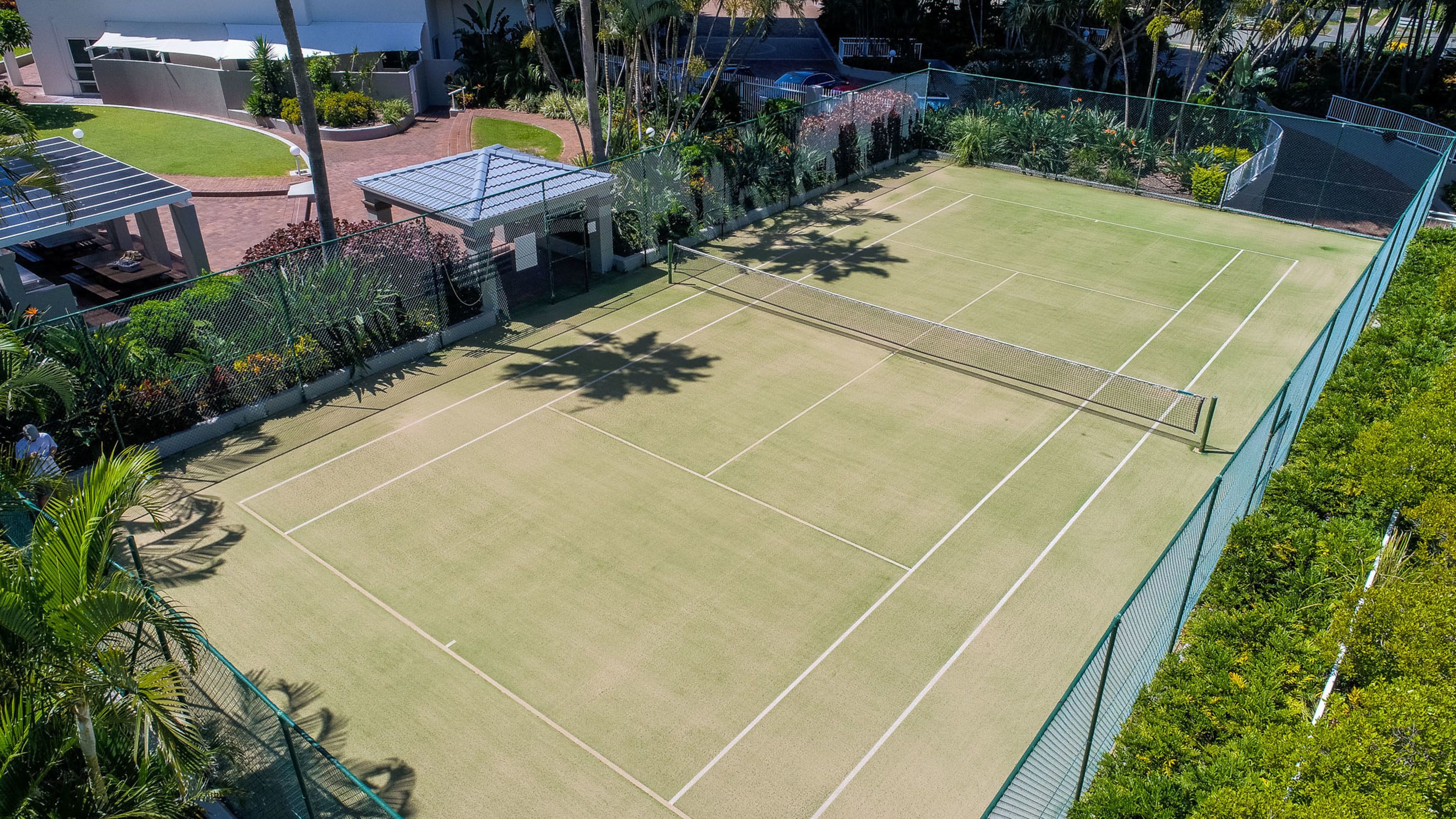 Tenniskenttä, jossa on verkko, ja jonka ympärillä on aita sekä kasvillisuutta.