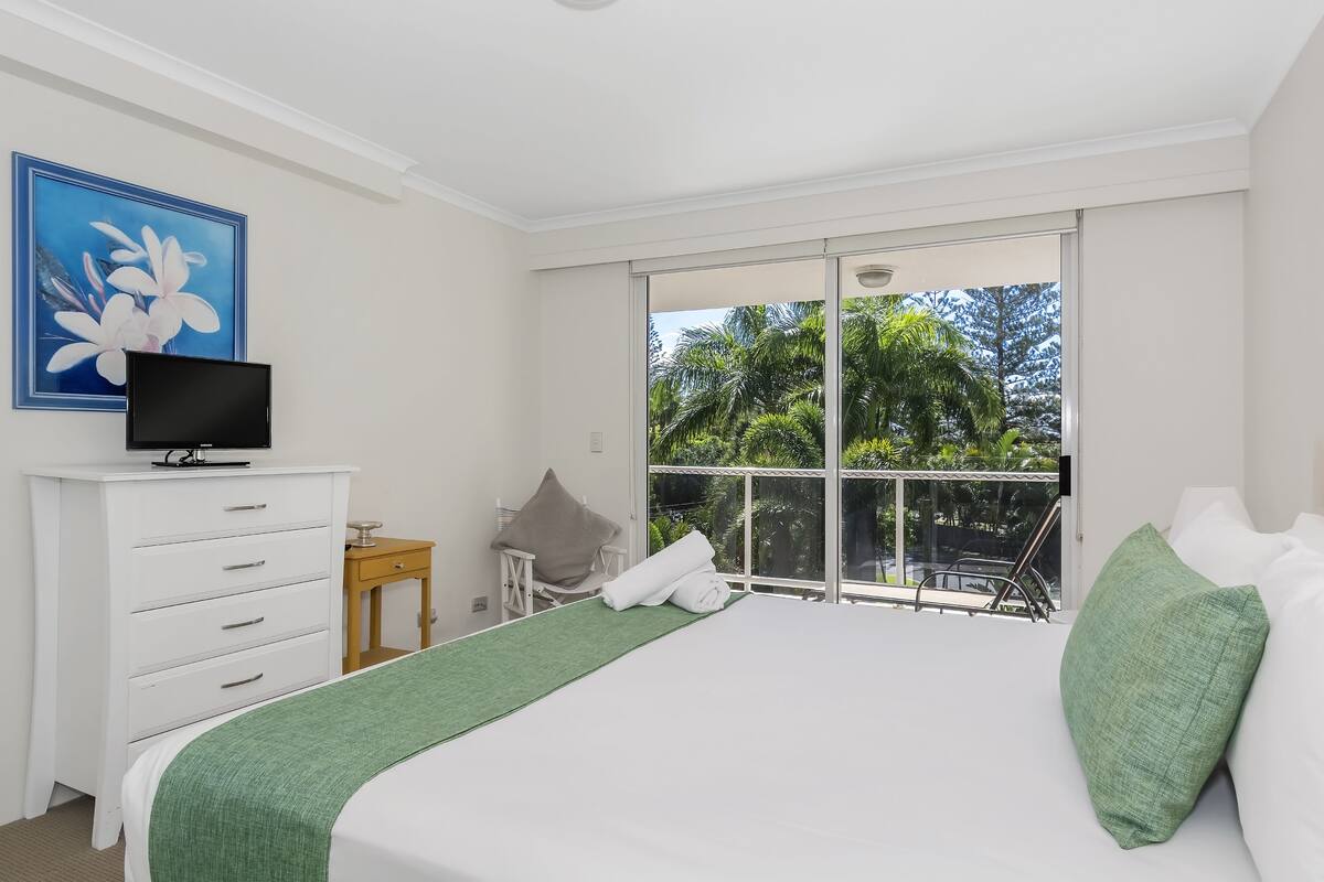 A bedroom with a bed, a TV on a dresser, a small table with a chair, and a large window with a view of trees.