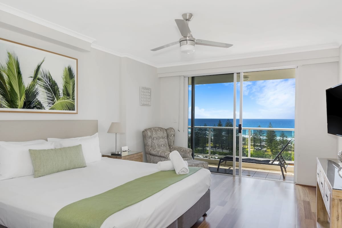 A hotel room with a bed, a chair, a TV, and a balcony with a view of the ocean.