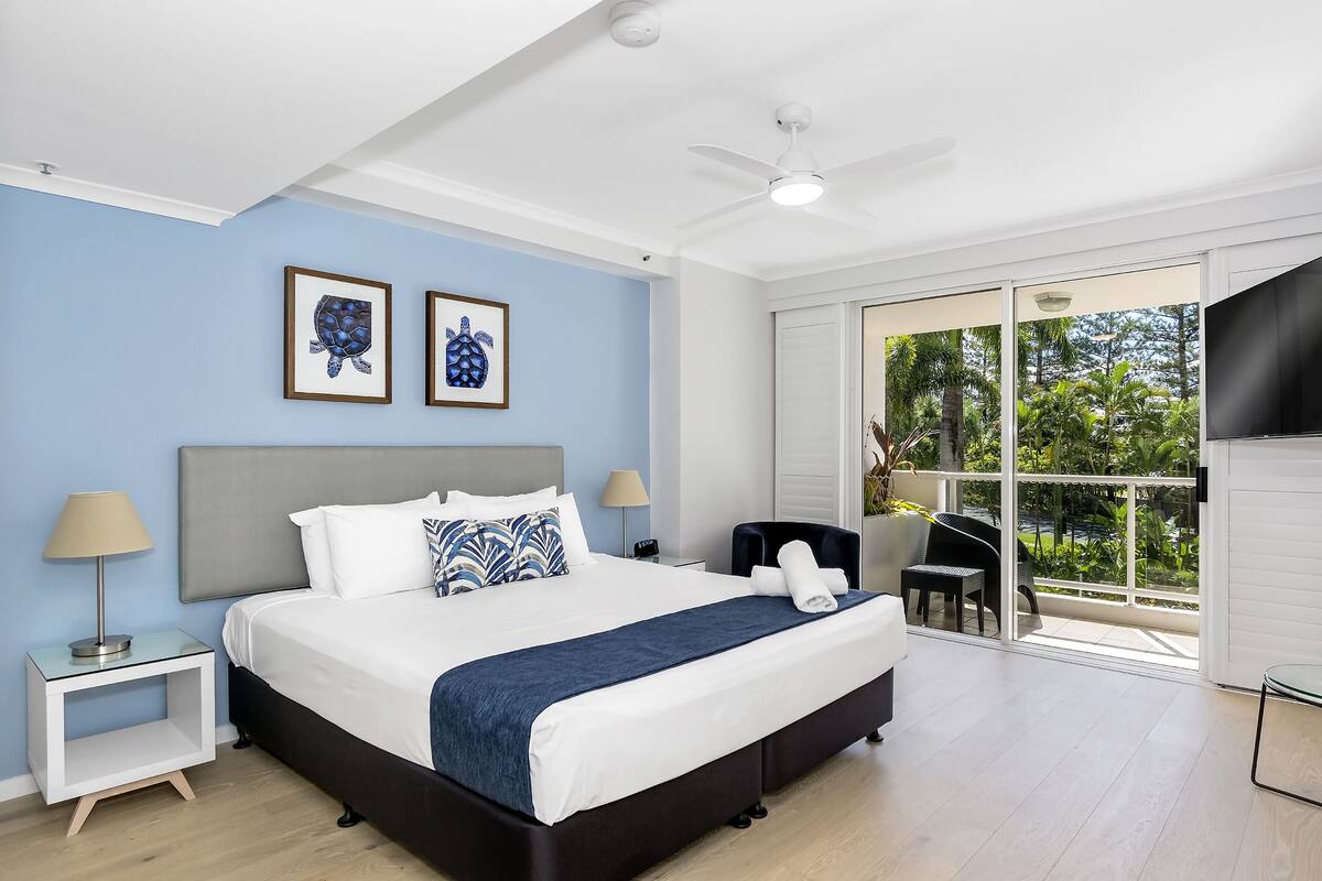A modern hotel room with a large bed, a ceiling fan, and a sliding glass door leading to a balcony with a view of greenery.