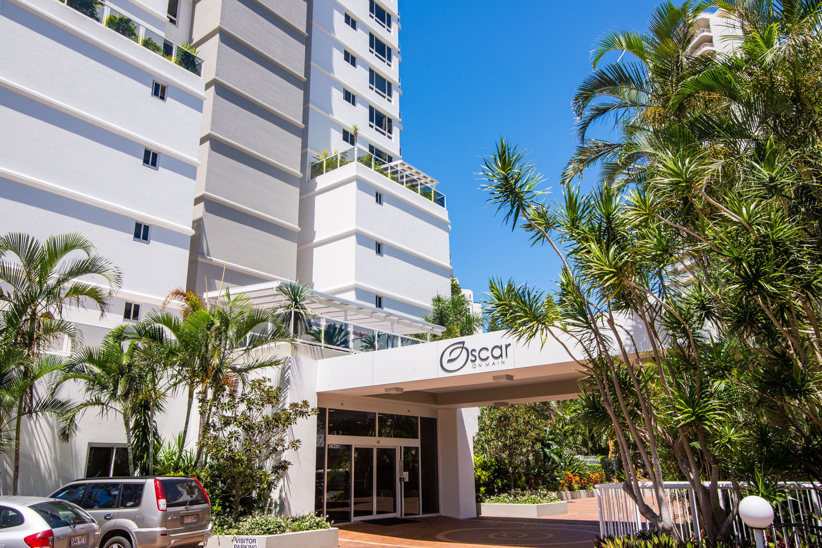 A modern hotel entrance with a white facade, palm trees, and a clear blue sky.