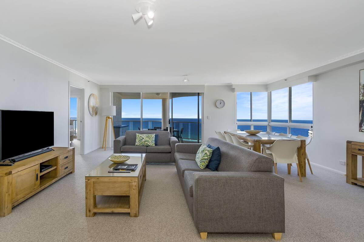 A modern living room with a large flat-screen TV, a wooden coffee table, and a gray sofa. There is a dining area with a table and chairs, and large windows offering a view of the ocean.