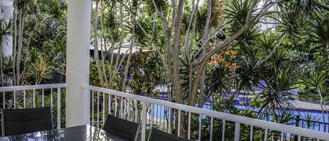 A balcony with a table and chairs, surrounded by lush tropical plants and a view of a pool.