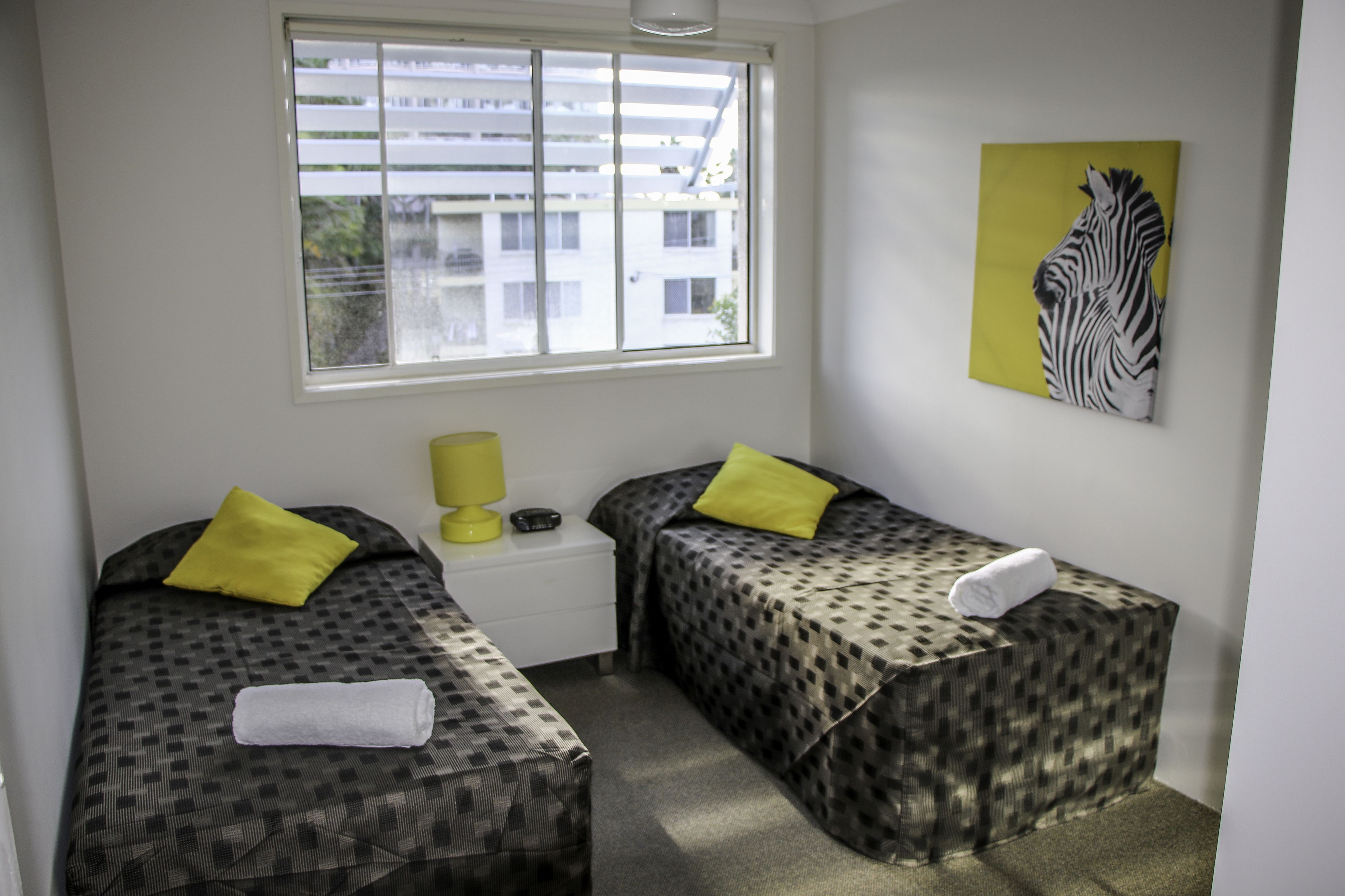 Una habitación pequeña y limpia con dos camas, cada una con una colcha estampada y un almohadón amarillo. Hay una ventana que proporciona luz natural y una obra de arte con diseño de cebra en la pared.