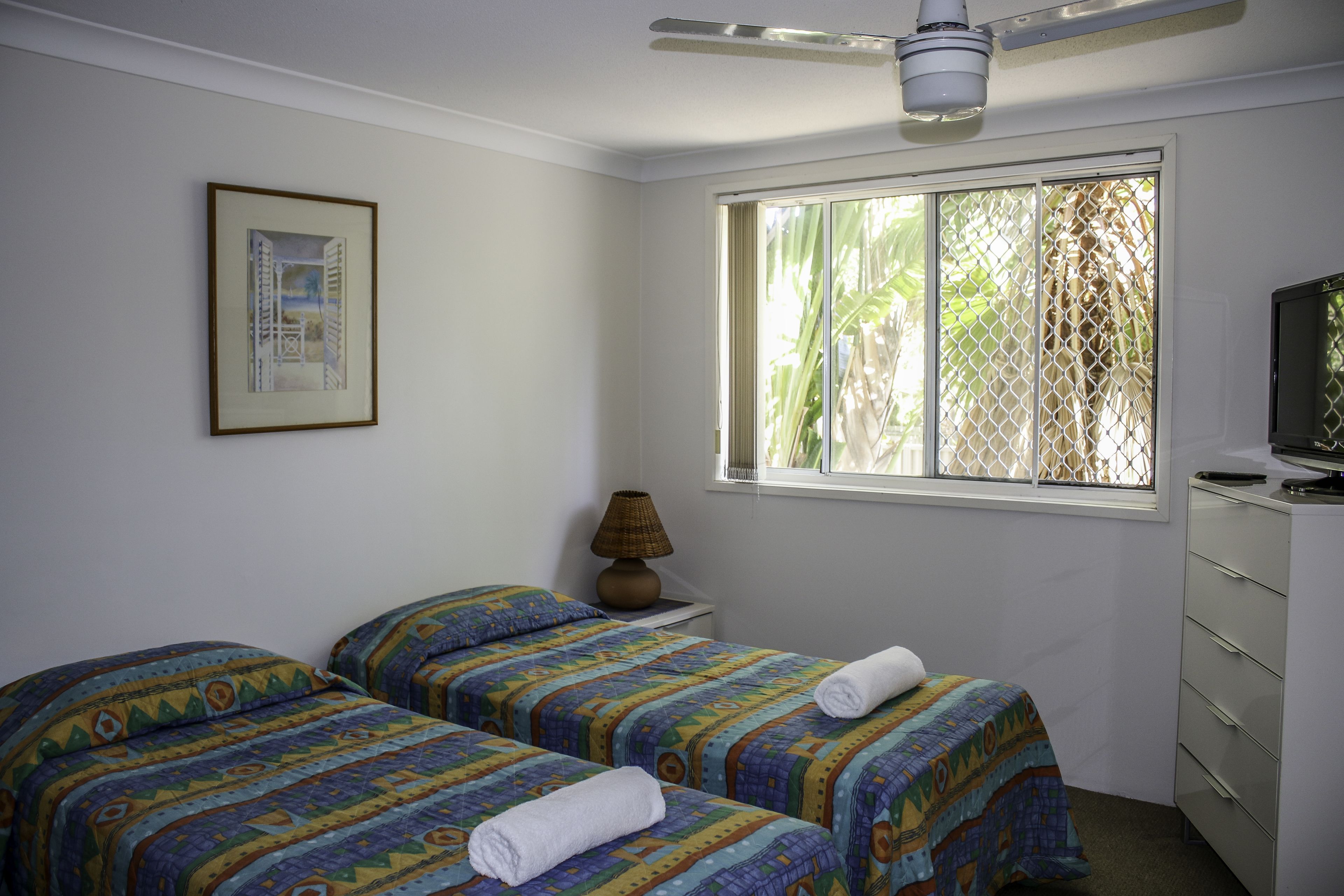 A bedroom with two beds, a window with a view of greenery, a ceiling fan, and a television on a dresser.