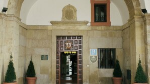 A stone entrance with a large archway, a door with a coat of arms, and two potted evergreen trees. - Parador de Santo Domingo Bernardo de Fresneda (Santo Domingo de la Calzada)