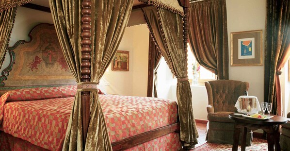 Una cama con dosel con un edredón rojo estampado, un cabecero de madera con un diseño pintado, y una silla con una mesa y dos copas de vino.