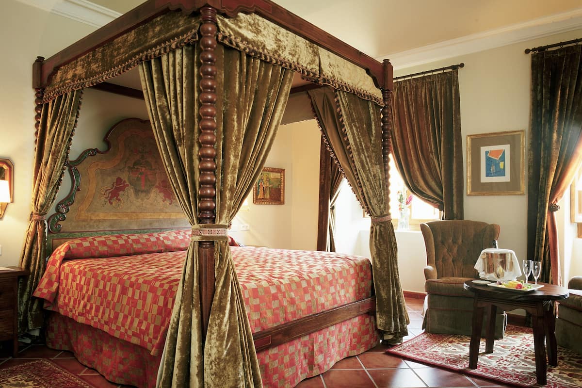 A four-poster canopy bed with a red and beige patterned bedspread, a wooden headboard with a floral design, and a red and beige patterned rug on a tiled floor.