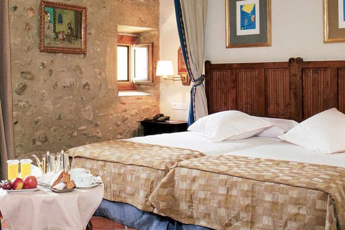 A double bed with a wooden headboard, a bedside table with a lamp, and a breakfast setup with fruits and pastries.
