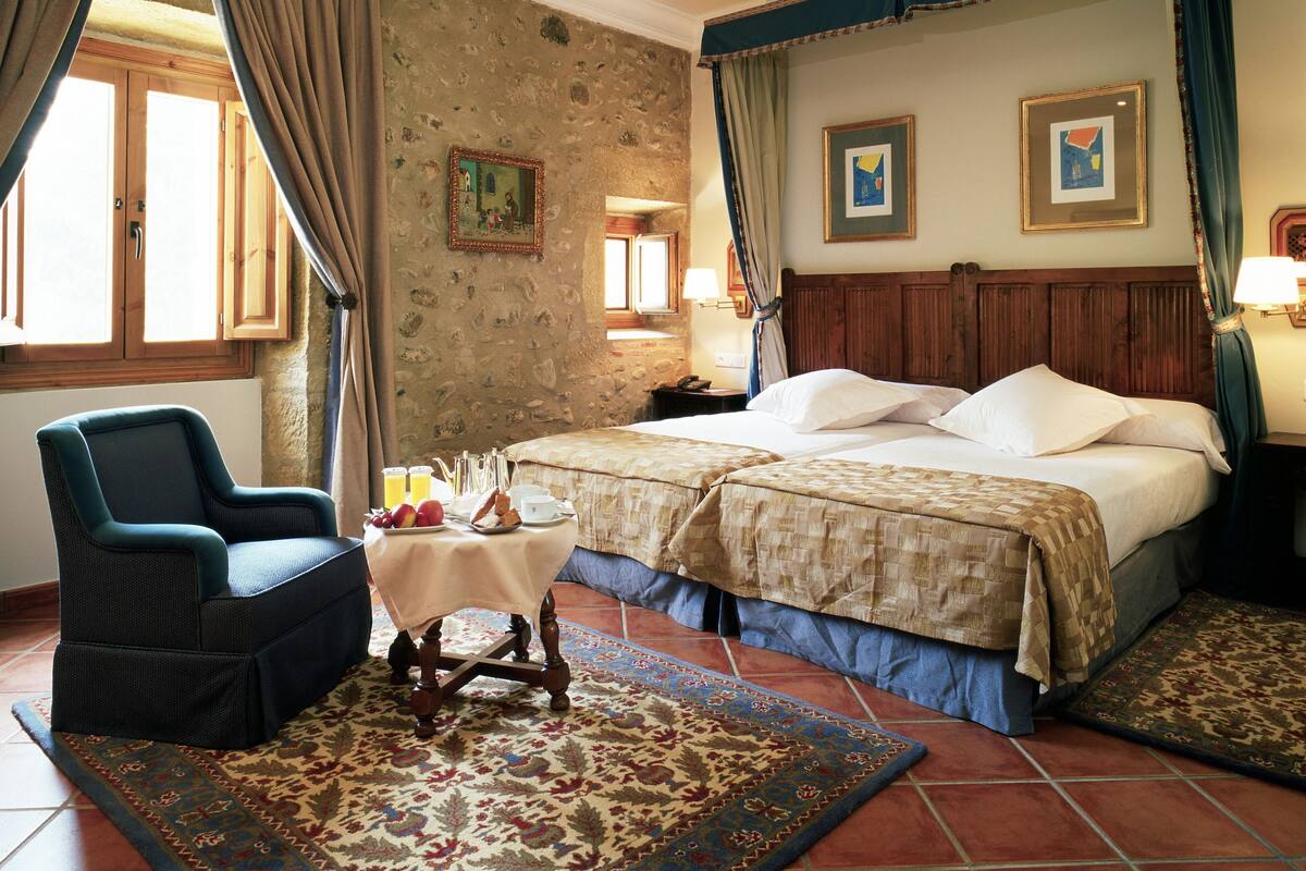 A bedroom with a large bed, a chair, a small table with breakfast items, and a patterned rug.