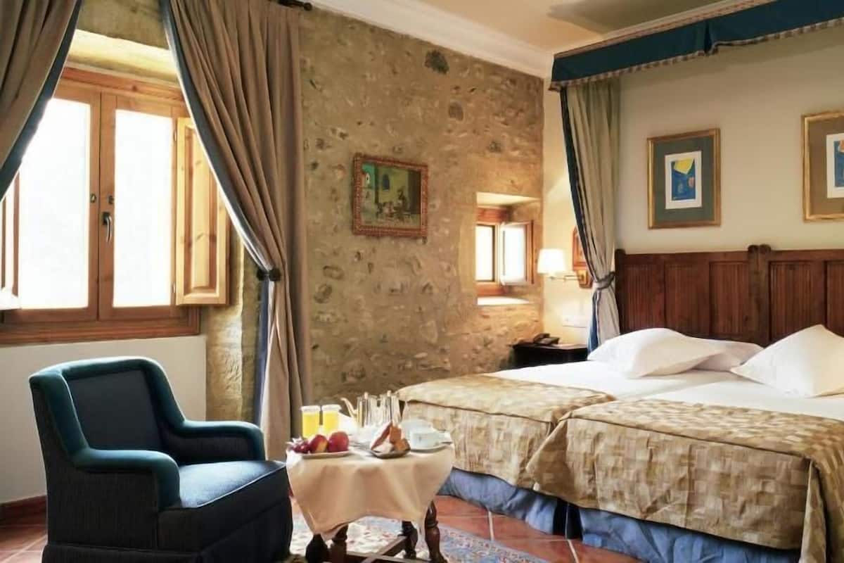 A room with a bed, a chair, a small table with fruits and a bottle, and a stone wall.