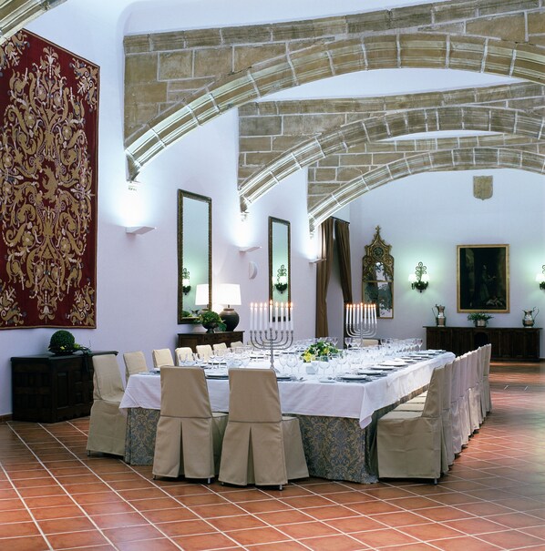 A formal dining room with a long table set for a meal, stone arches, and a large tapestry on the wall.