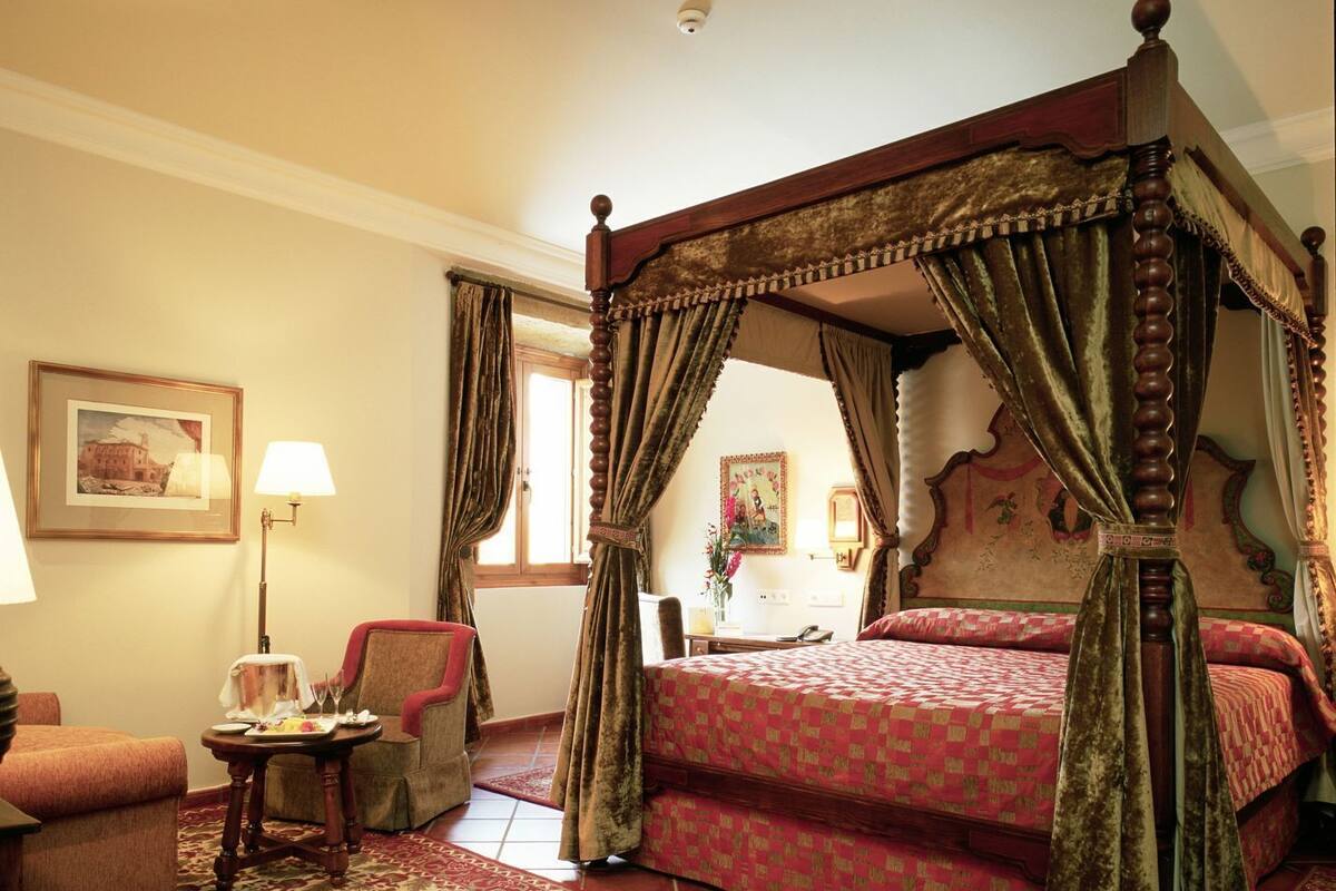 A four-poster bed with a canopy, a chair, a small table with a lamp, and a framed picture on the wall.