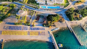 Vue aérienne d’un complexe hôtelier doté d’une piscine, d’une plage avec des parasols et d’une route menant aux bâtiments.