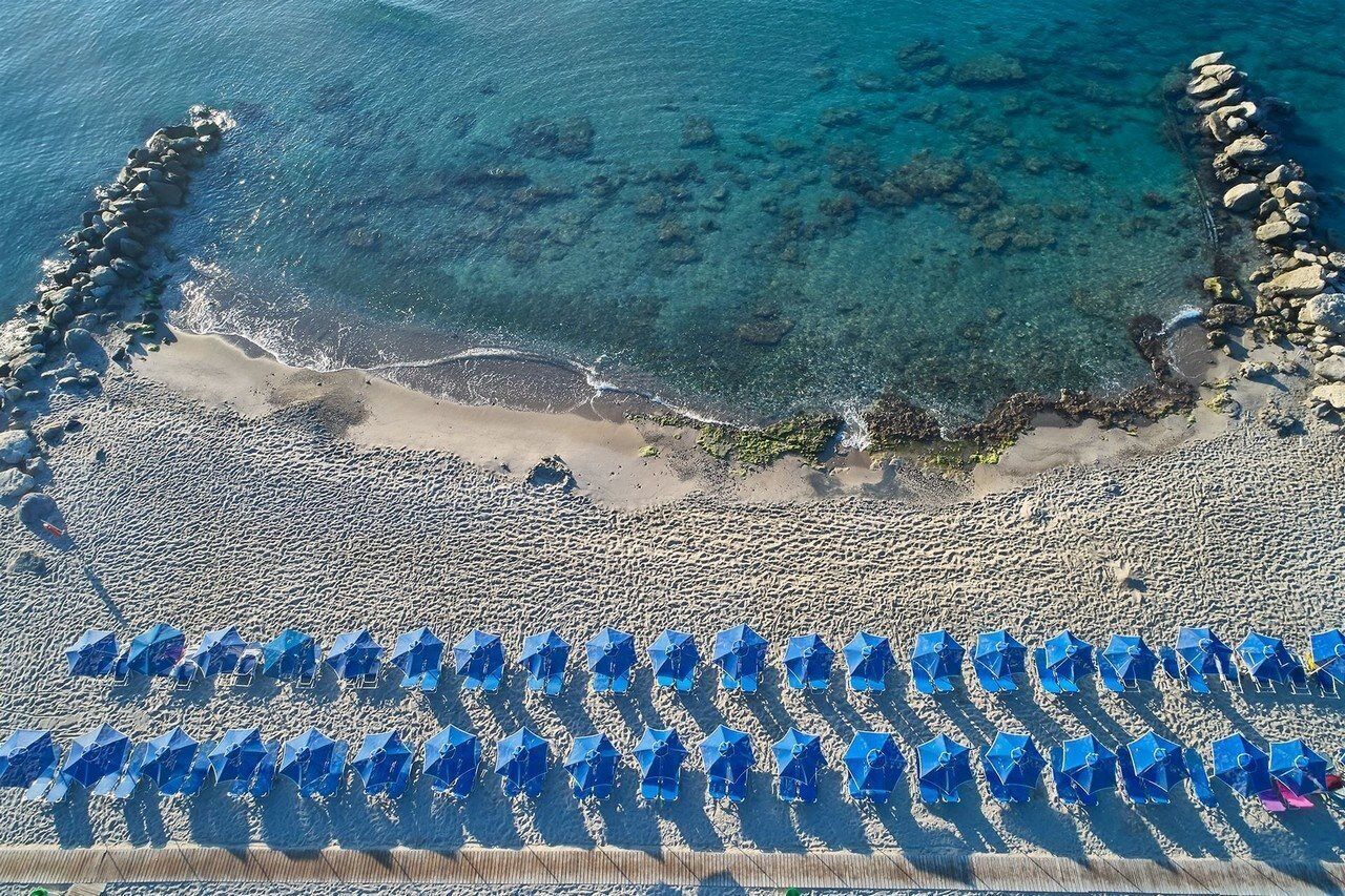 Eine Vogelperspektive zeigt einen Strand mit klarem, blauem Wasser, einer felsigen Küste und Reihen von blauen Sonnenschirmen.