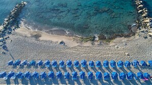 Une vue aérienne d’une plage avec une eau d’un bleu limpide, un rivage rocheux et des rangées de parasols bleus.