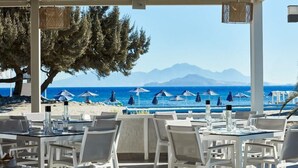 Un restaurant situé en bord de mer, avec une terrasse, des tables et des parasols, offrant une vue sur la mer et les montagnes.