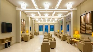A spacious hotel lobby with a modern design, featuring a central seating area with sofas and armchairs, a flat-screen TV mounted on the wall, and a large window providing a view of the cityscape. - Lagas Aegean Village (Kos Town)