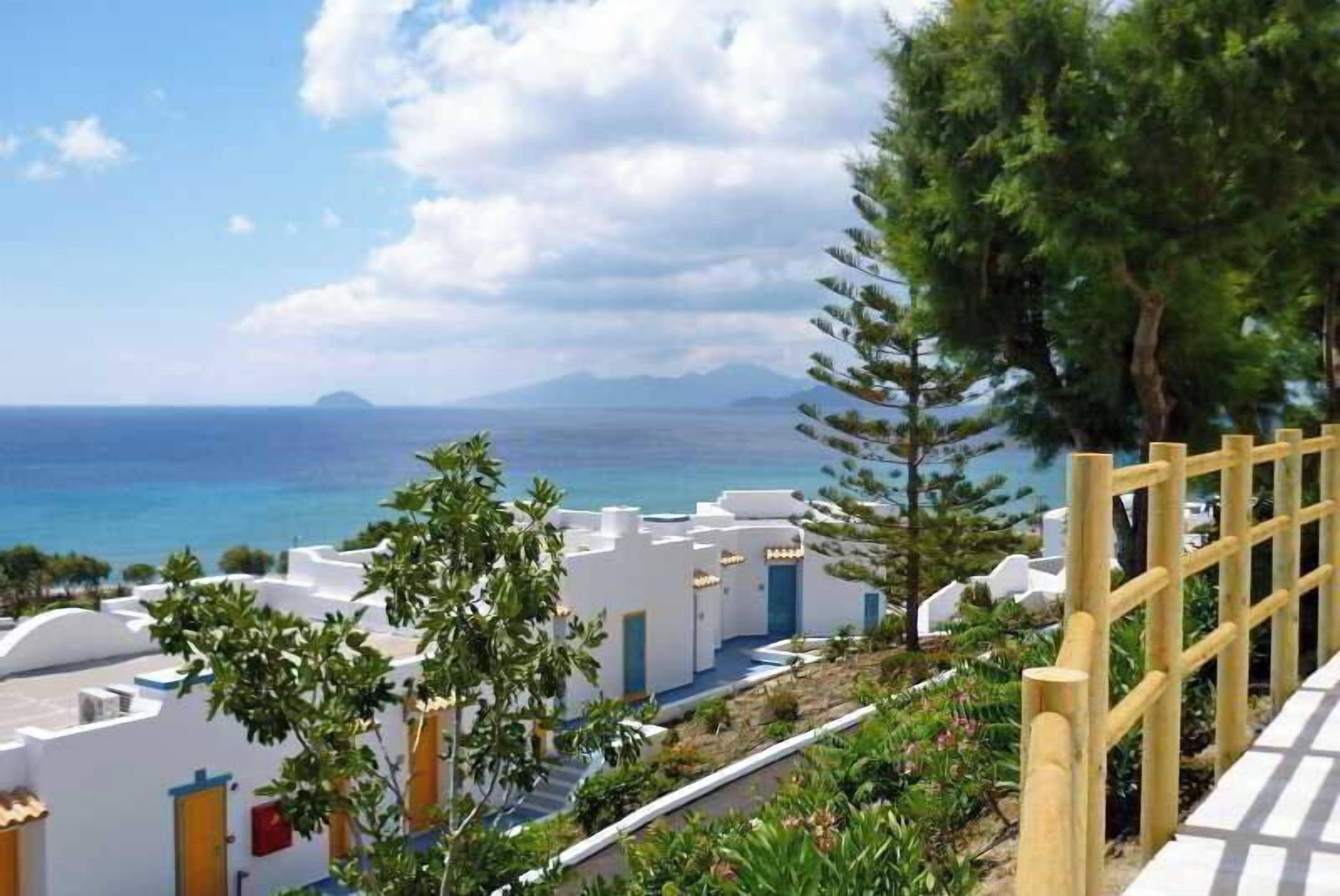 A coastal hotel with white buildings, a wooden fence, and a view of the sea and mountains.