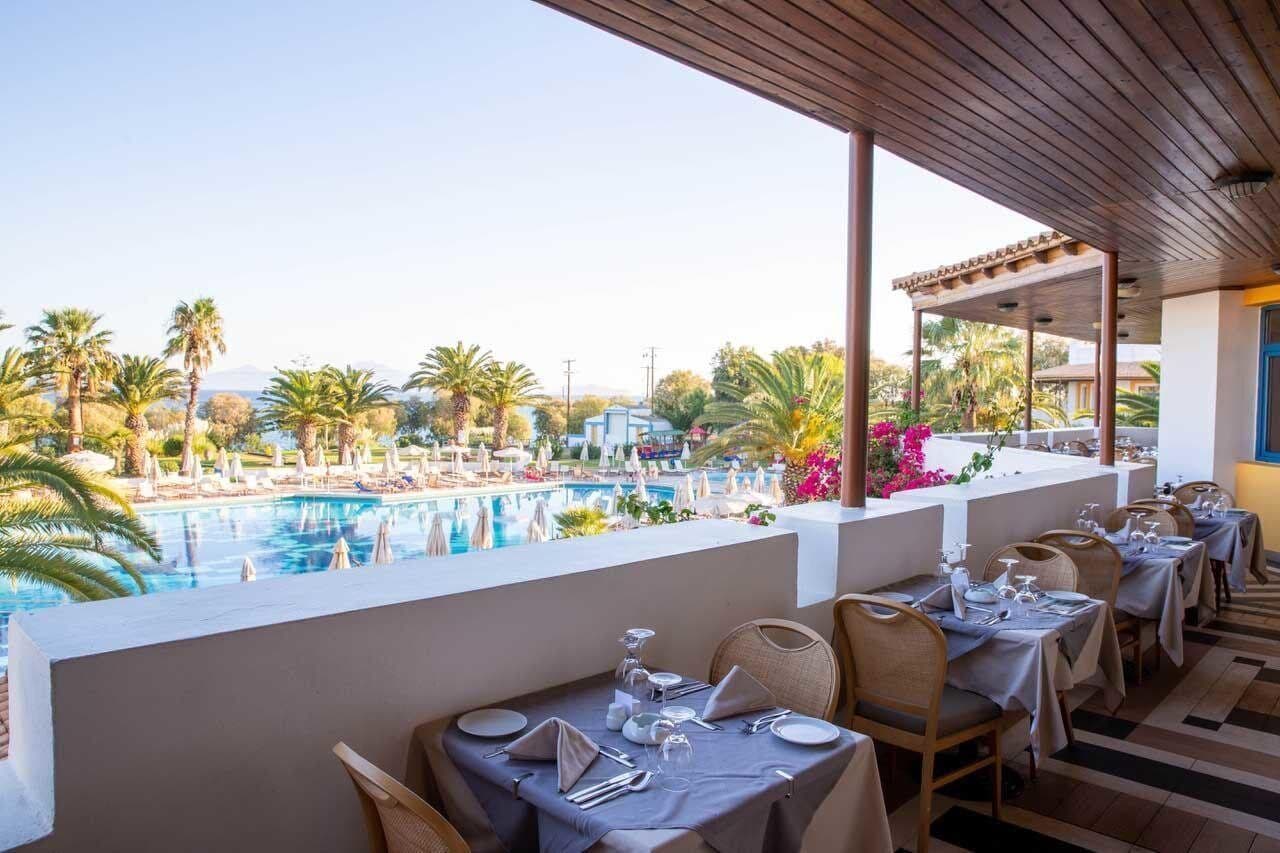Un restaurante con terraza al aire libre que da a una piscina, rodeado de palmeras y un cielo despejado.