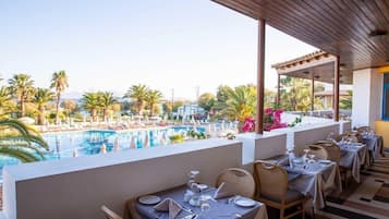 Un restaurante con terraza al aire libre que da a una piscina, rodeado de palmeras y un cielo despejado.