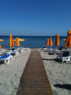 A beach with a wooden walkway leading to the sea, surrounded by sun loungers and orange umbrellas. - Lagas Aegean Village (Kos Town)