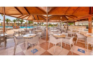 An outdoor dining area with tables and chairs, a pool, and umbrellas in the background. - Lagas Aegean Village (Kos Town)