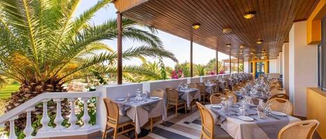 An outdoor dining area with multiple tables and chairs, a covered roof, and a view of palm trees.