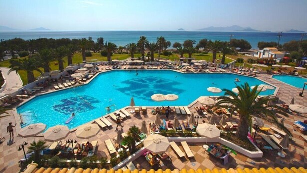 A large outdoor swimming pool with multiple sun loungers and umbrellas, surrounded by palm trees and a view of the sea. - Lagas Aegean Village (Kos Town)